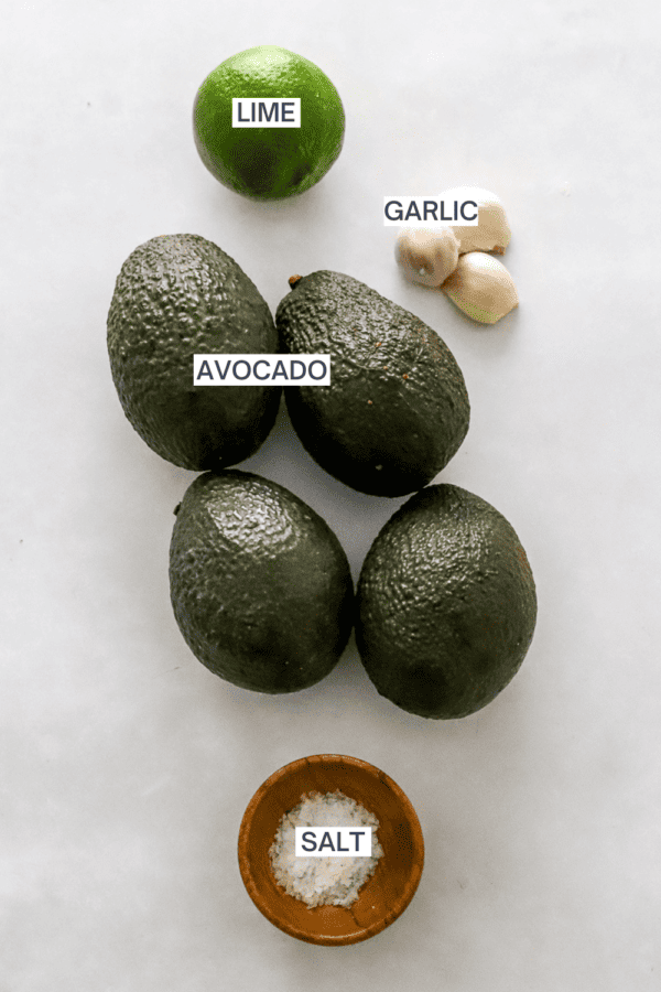 Avocados, garlic cloves, lime and salt in a small wood bowl with labels over each ingredient.