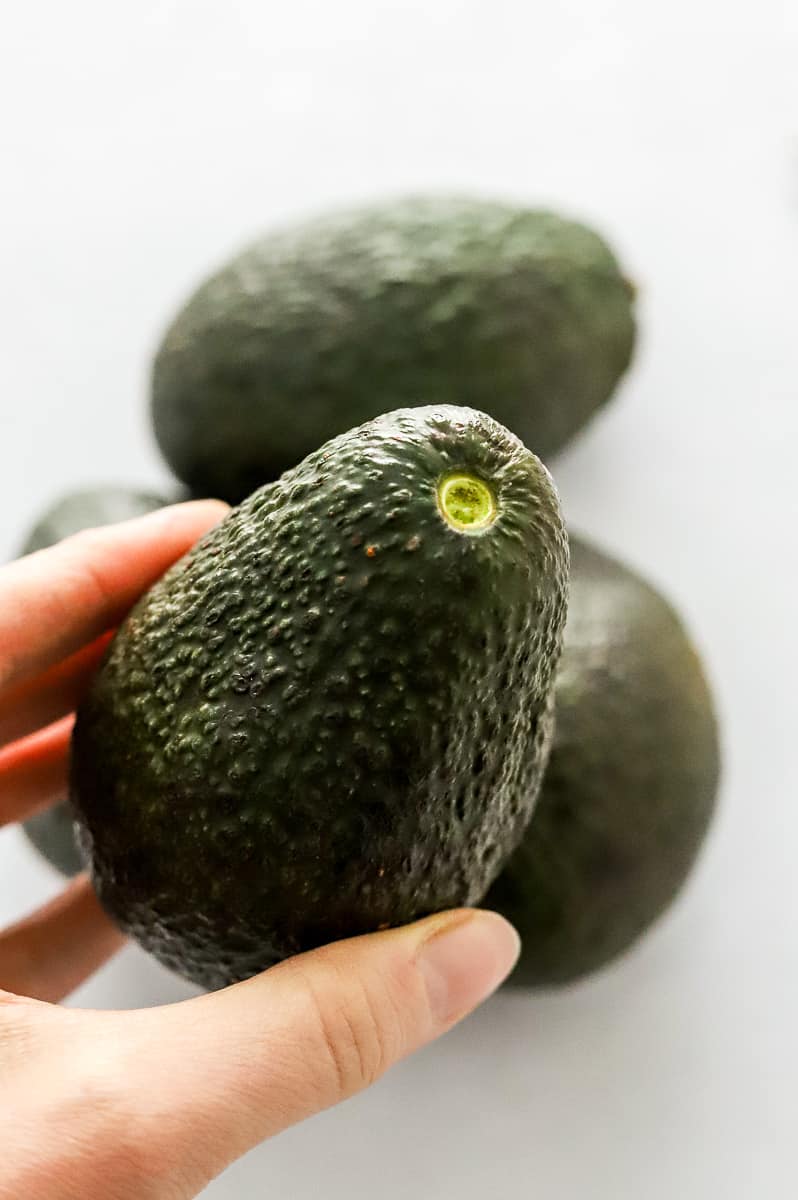 Hand holding a whole avocado with more avocados behind it.