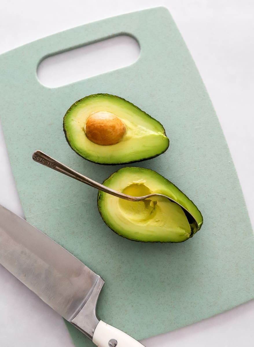 Sliced avocado on a blue cutting board with a spoon in one half of the avocado with a white knife next to it.