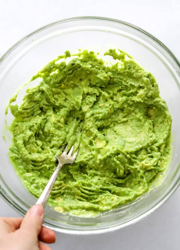 Hand using a fork to mash avocados in a glass bowl.