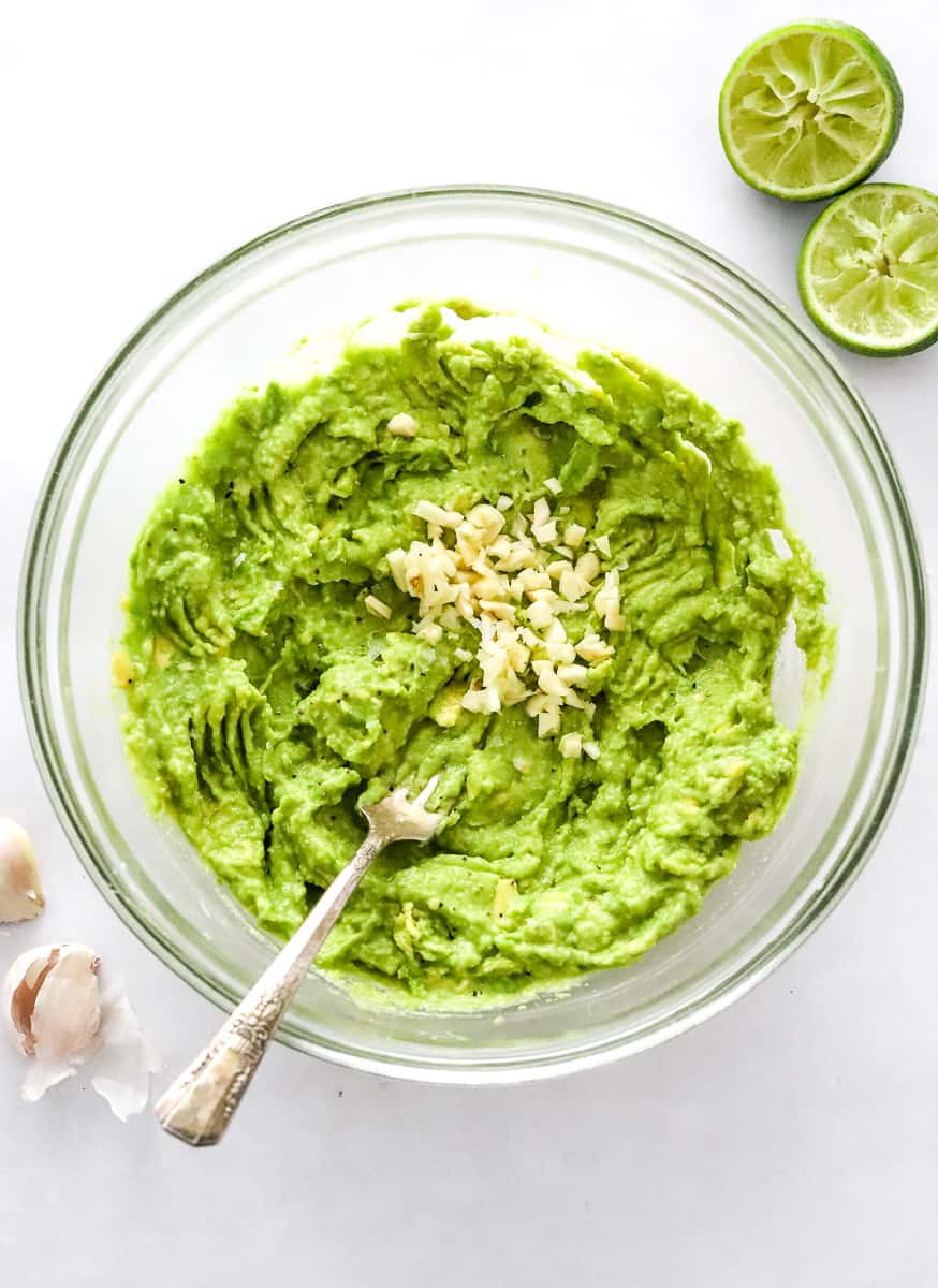 Mashed avocados in a glass bowl with minced garlic on it and squeezed limes behind it.