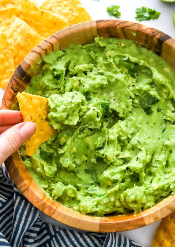 Hand dipping a tortilla chip in a brown bowl filled with 4 ingredient guacamole with more chips behind it and blue and white linen in front of it.