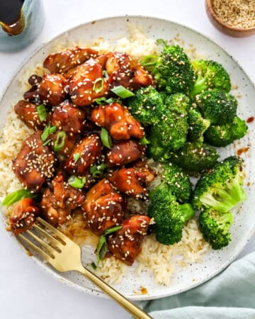 Plate of cooked air fryer chicken teriyaki on top of rice with broccoli next to it and more teriyaki sauce and sesame seeds behind it with a gold fork on the plate.