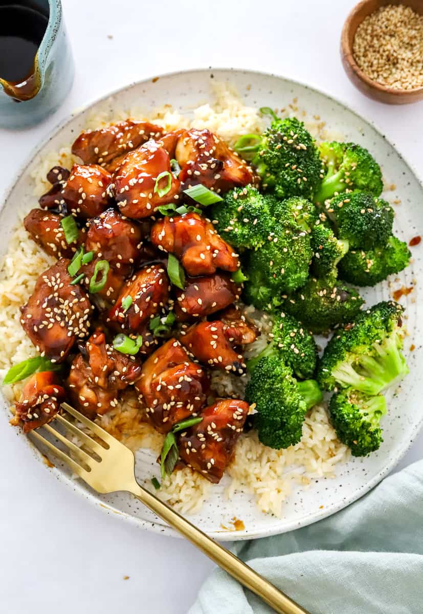Plate of cooked air fryer chicken teriyaki on top of rice with broccoli next to it and more teriyaki sauce and sesame seeds behind it with a gold fork on the plate.