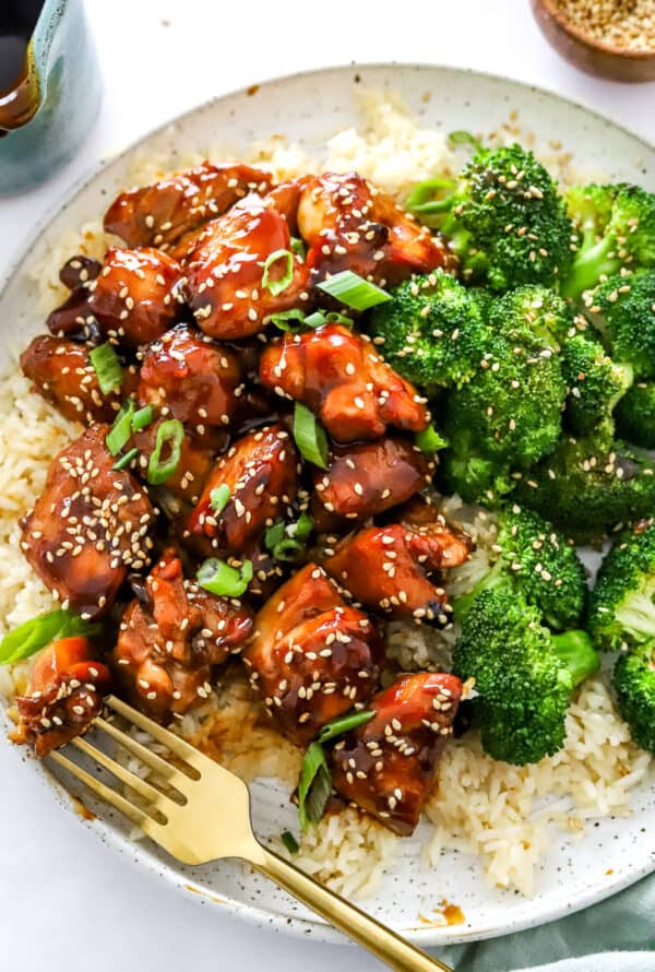 Chicken teriyaki with broccoli on top of rice on a plate with a fork topped with sesame seeds with more sesame seeds and sauce behind it.