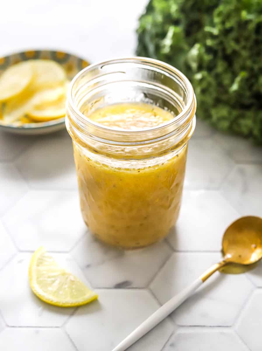Lemon kale salad dressing in a mason jar with a spoon with dressing on it and a lemon slice in front of it and a bunch of kale and sliced lemons behind it.