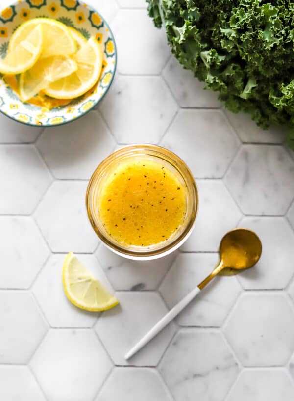 Jar filled with lemon dressing with a spoon and slice of lemon in front of it and curly kale and a bowl of sliced lemons behind it.