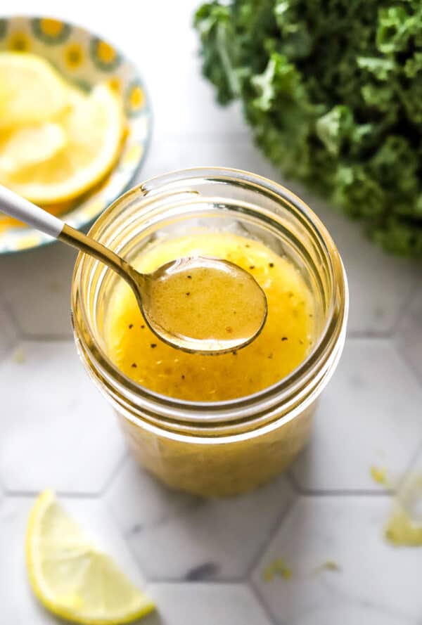 Spoon lifting out of a jar of salad dressing with dressing on it with a bunch of kale and lemons around it.