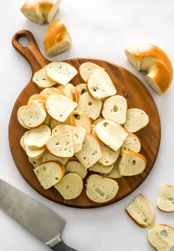 Air fryer bagel chips sliced on a round cutting board with more cut bagels behind it and a kitchen knife in front of it.