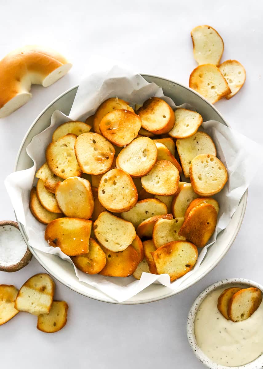 Round bowl lined with parchment paper filled with golden brown cooked bagel chips with more chips around it. 