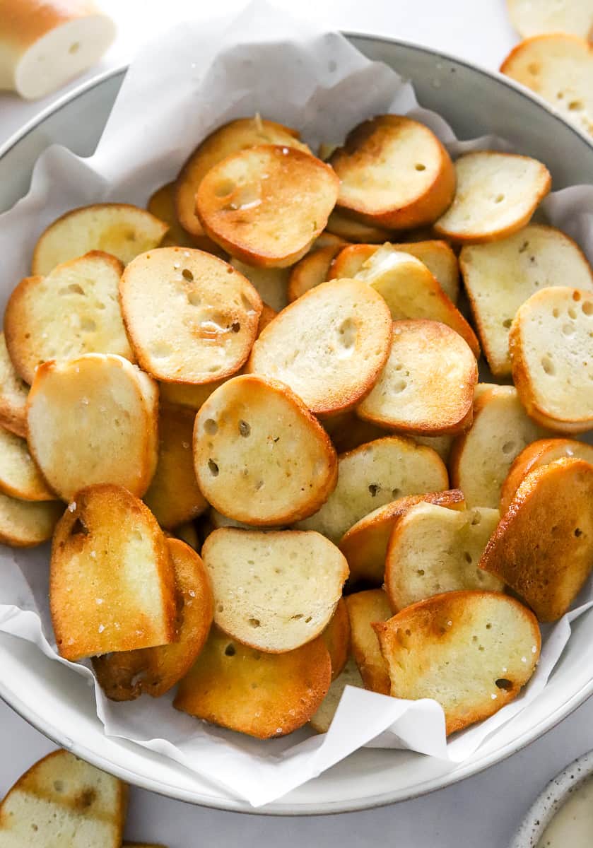Cooked golden bagel chips in a parchment lined bowl with more chips around it.