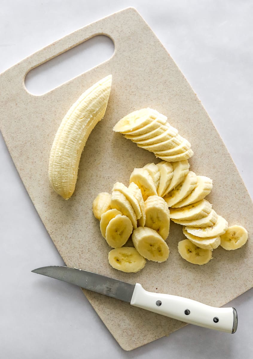 Sliced banana on a light brown cutting board with a white handled knife on the cutting board next to the banana.