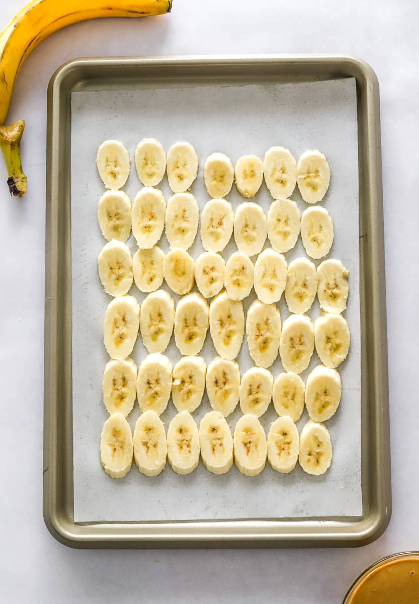 Sliced banana pieces in rows on a parchment lined baking sheet with a banana peel behind it and a bowl of peanut butter in front of it.