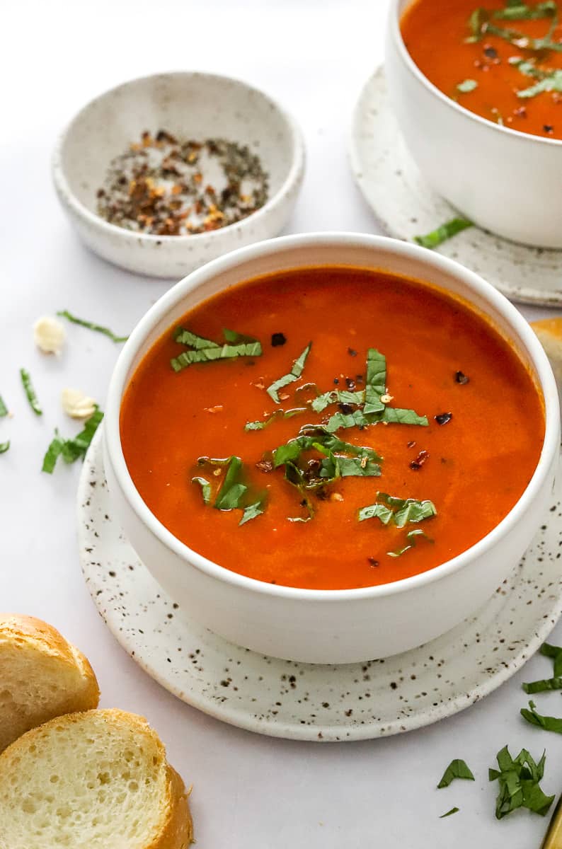 Two bowls of tomato soup topped with chopped basil with a bowl of red pepper flakes next to it. 