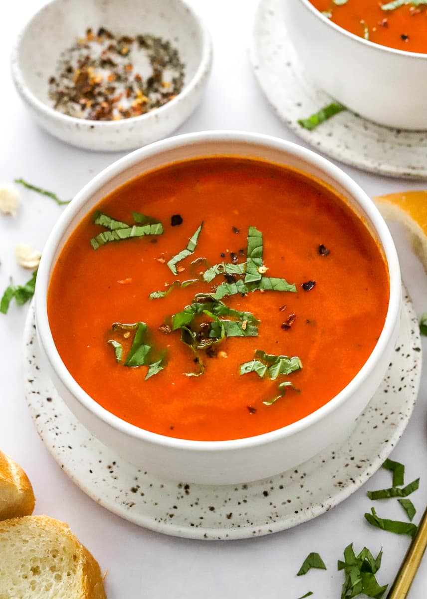 White bowl filled with vibrant tomato soup with fresh herbs on top with another bowl of soup behind it and slices of bread in front of it. 