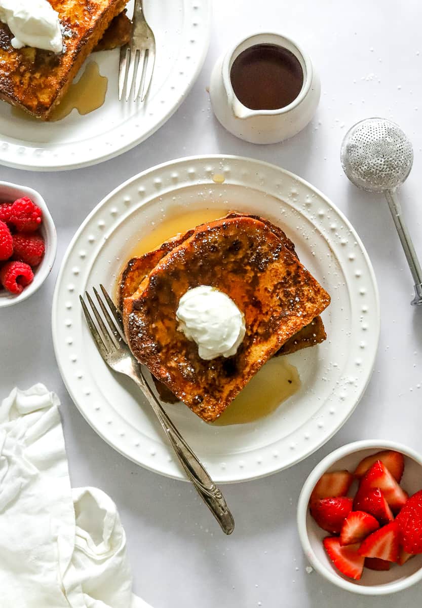 Two plates of French toast topped with yogurt, and syrup with berries in white bowls around it. 