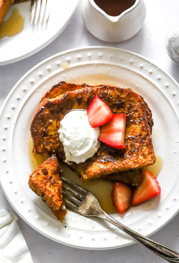 White plate with 2 slices of protein French toast topped with yogurt, strawberries and syrup with a fork on the plate.