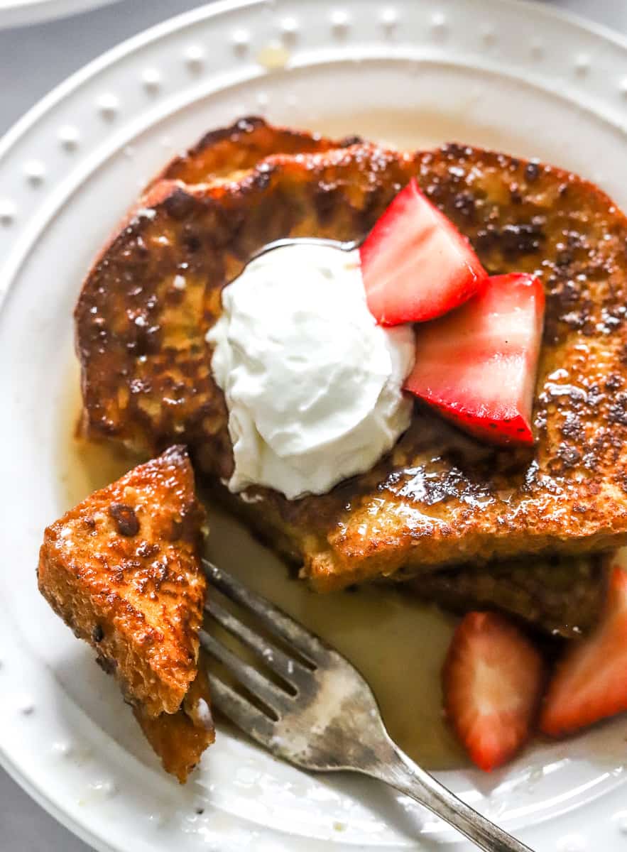 Two pieces of golden French toast covered in syrup with a dollop of yogurt and sliced strawberries on it with a fork holding and piece of the toast. 