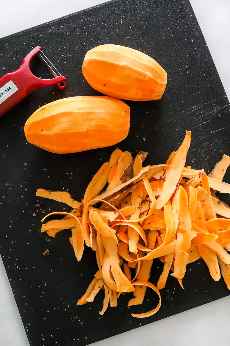 Two peeled sweet potatoes on a black cutting board with the peels in front of them and a red peeler behind them.