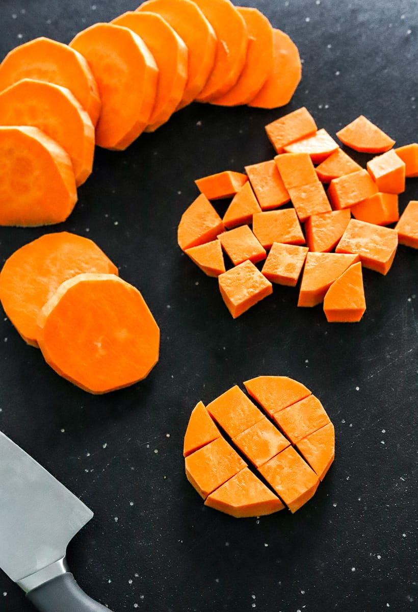 Peeled and cubed sweet potatoes on a black cutting board with some round cut sweet potatoes behind them on the board. 