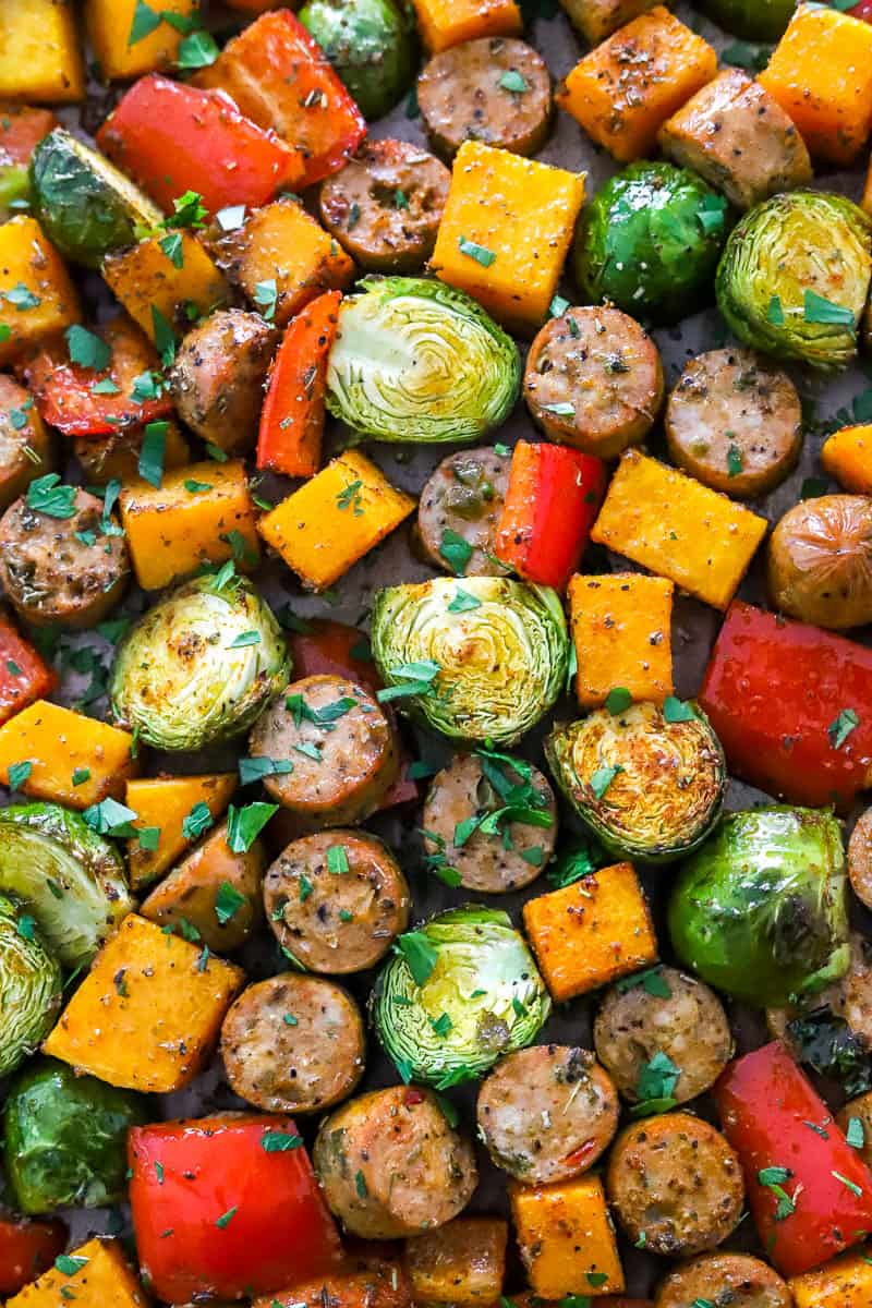 Close up shot of roasted squash, Brussels, peppers and sliced sausage on a baking sheet topped with chopped herbs.