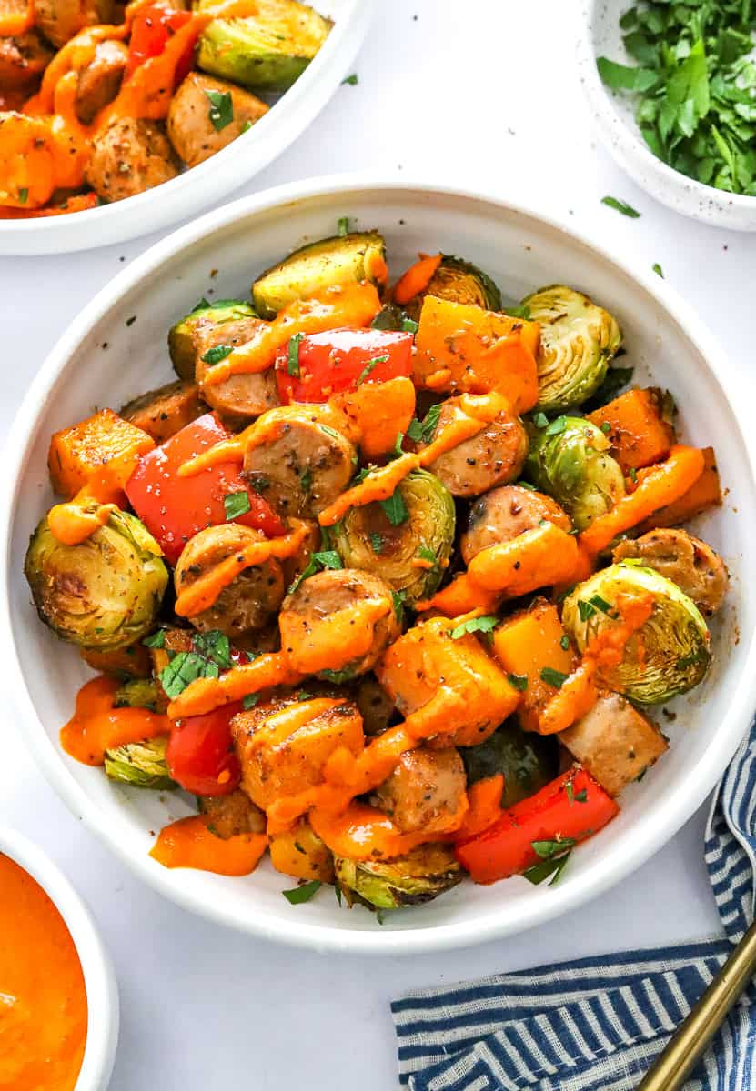 Round bow filled with chopped roasted veggies and sausage with orange sauce drizzled over it with another bowl of it and herbs behind it.