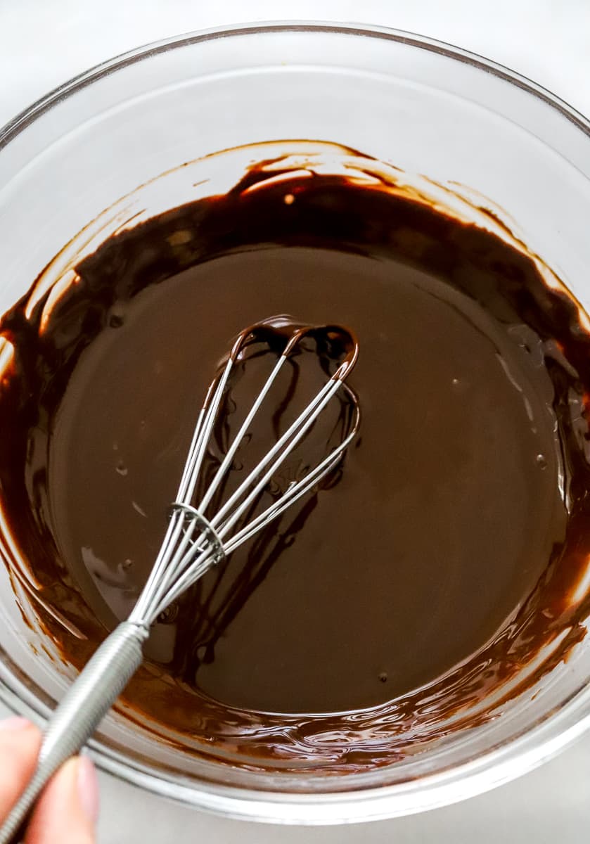 Melted chocolate in a glass mixing bow with a hand mixing it with a metal whisk.