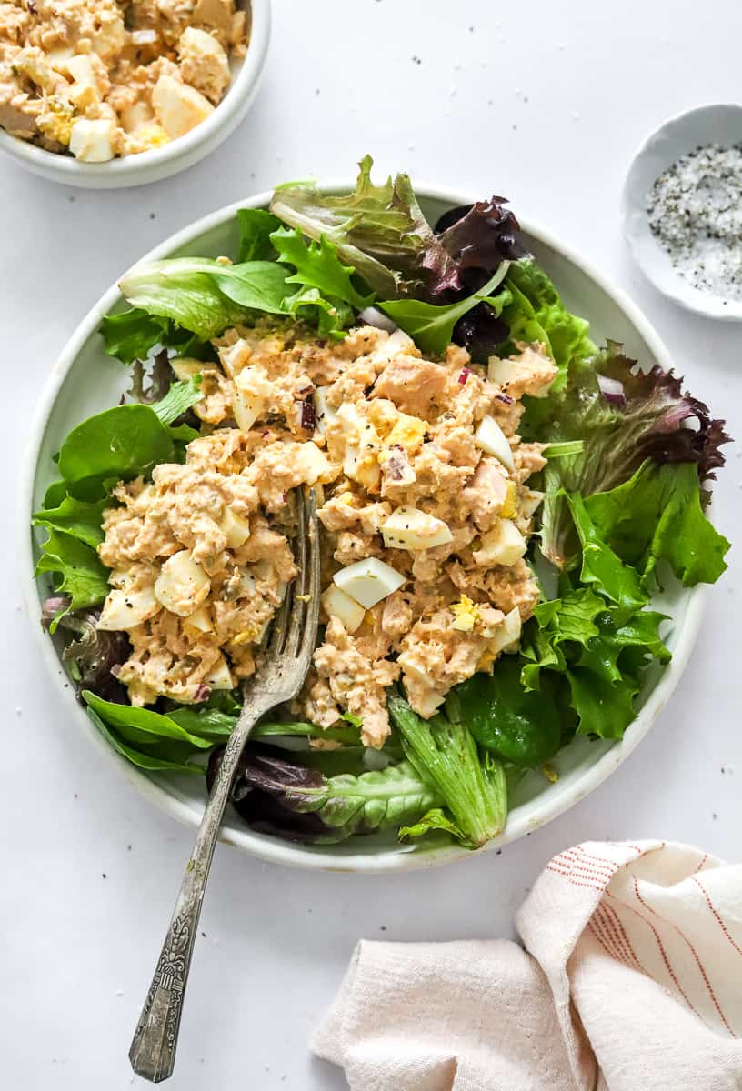 Tuna and egg salad over mixed greens on a plate with a fork on it with more tuna salad in a bowl behind it. 