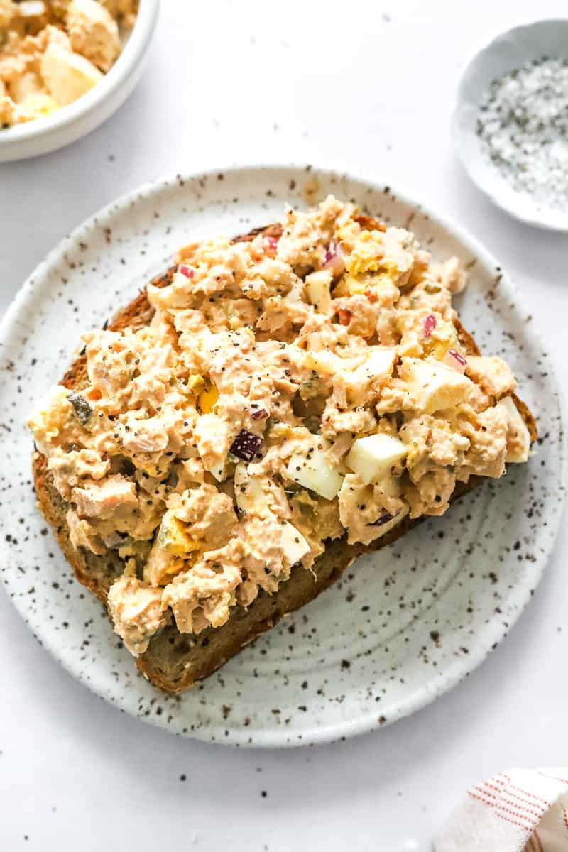 Tuna egg salad spread on a whole grain bread on a white and brown specked plate with more tuna salad in a bowl behind it. 