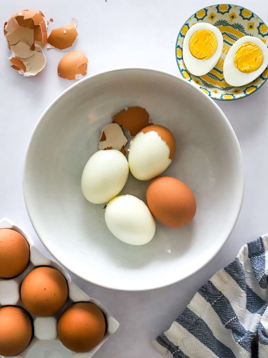 Hard boiled eggs in a white bowl with a few unpeeled with more in front of it and a sliced boiled egg and egg shells behind.
