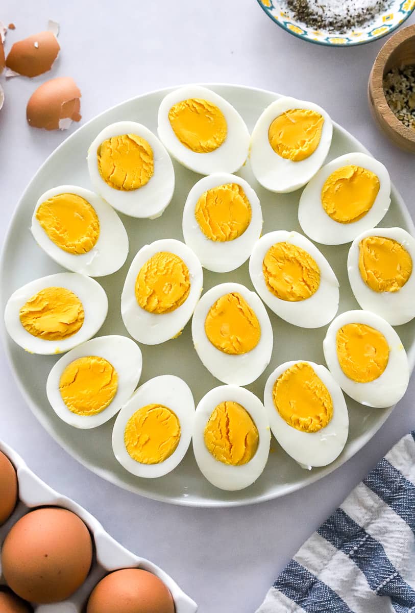 Sliced boiled eggs on a round plate with some whole eggs in front of it and egg shells and salt and pepper in a bowl behind it.
