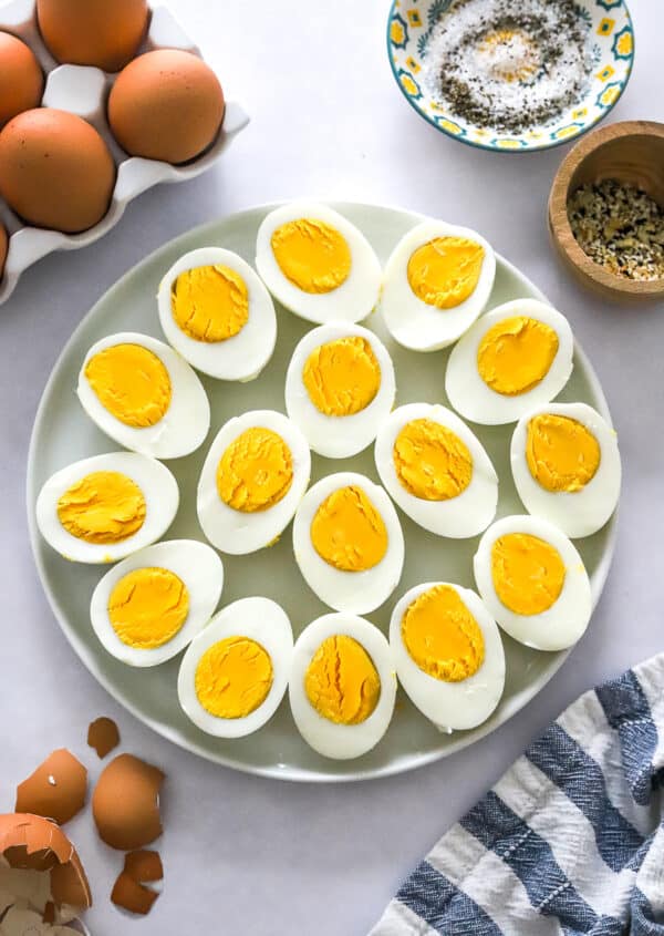 Place of sliced cooked whole hard boiled eggs with striped towel and egg shells I front of it and more eggs and bowl of seasoning behind it.
