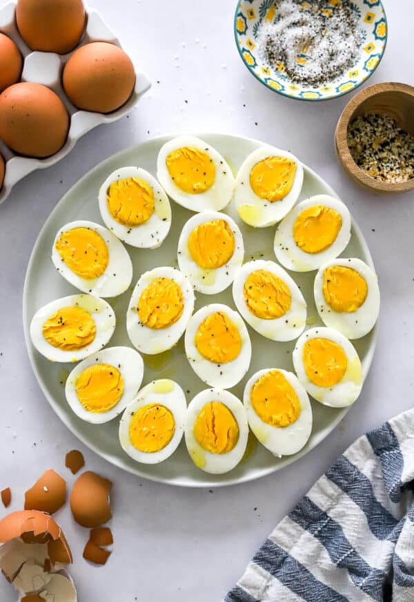 Instant pot hard boiled eggs on a round plate sliced in half with more eggs and spices in a bowl behind it and egg shells ad a striped towel in front of it.