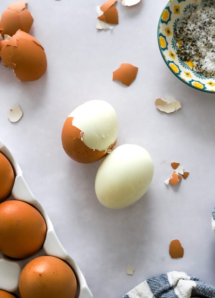 Half peeled hard boiled eggs with more eggs around it with a bowl of salt and pepper behind it and a blue and white striped towel in front of it.