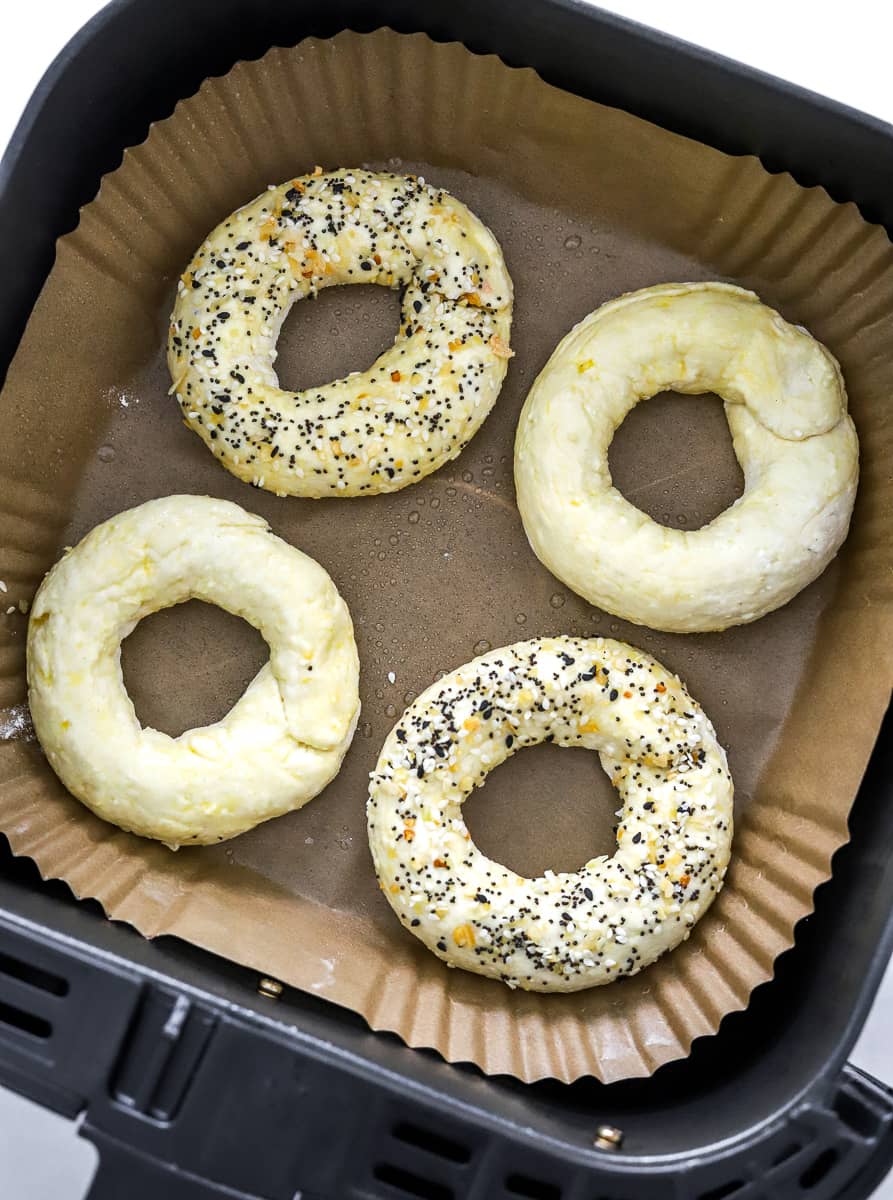 4 uncooked formed bagels in an air fryer basket lined with brown paper with everything seasoning on two of the bagels.