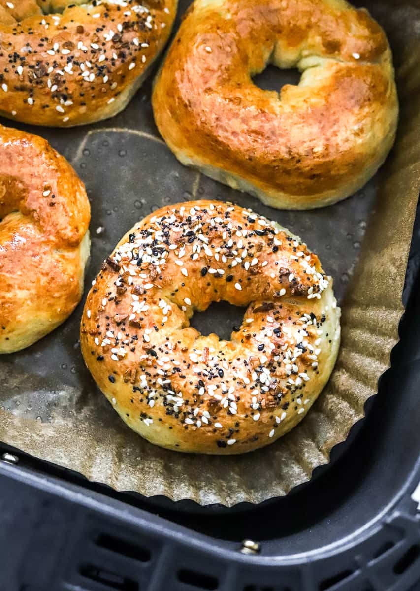 Everything bagel in an air fryer basket on top of brown parchment paper with more bagels behind it. 