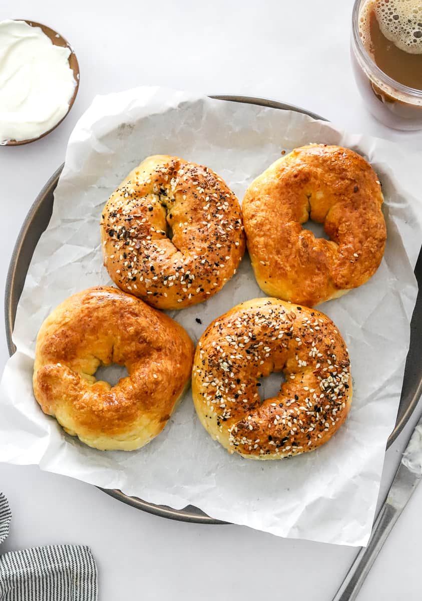 4 cooked protein bagels on a lined plate with a bowl of cream cheese and mug of coffee behind them and a knife and stripped towel in front of them.