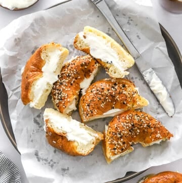 Lined plate with halves of cut bagels with cream cheese on them with more cream cheese and a coffee behind it.