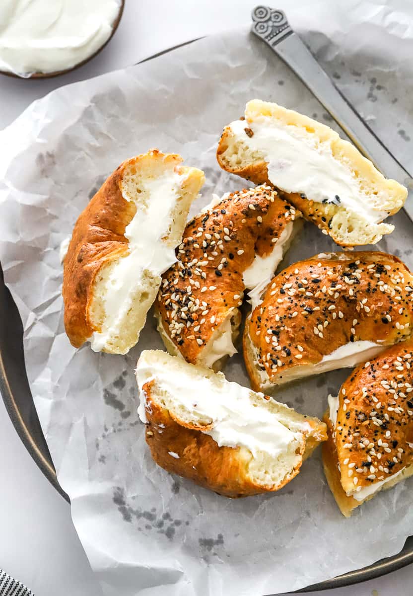 Halves of bagels with cream cheese on them on a parchment lined plate with a knife of the plate and more cream cheese in a bowl behind them.