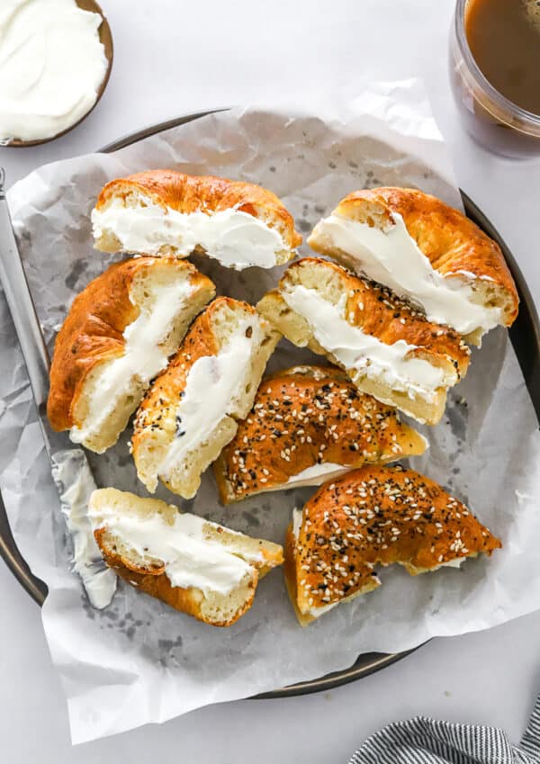 Plate of cream cheese bagel sandwiches cut in half with everything seasoning on some of them with more cream cheese and a coffee behind them.