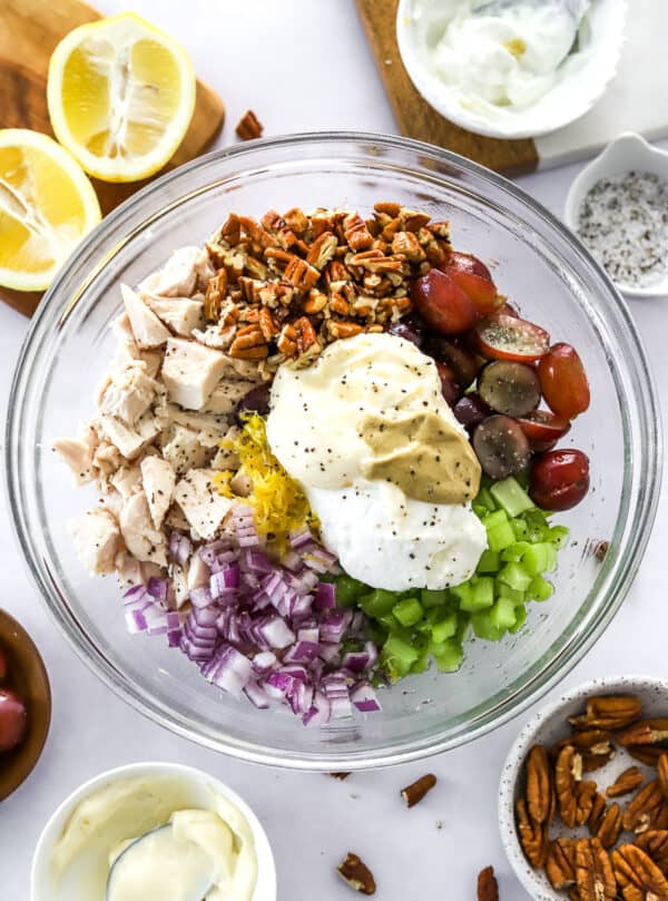 Round glass bowl filled with diced cooked chicken, chopped red onion, diced celery, chopped pecans, sliced grapes, mayo, yogurt, mustard with more of the ingredients around it.