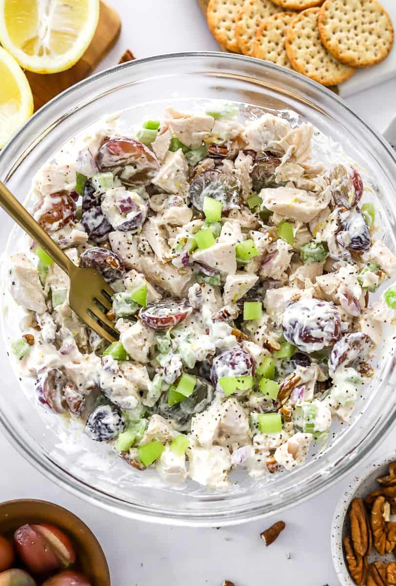 Mixed up chicken salad in a round glass bowl with a for in the bowl with pecans and grapes in front of it and a sliced lemon and crackers behind it.