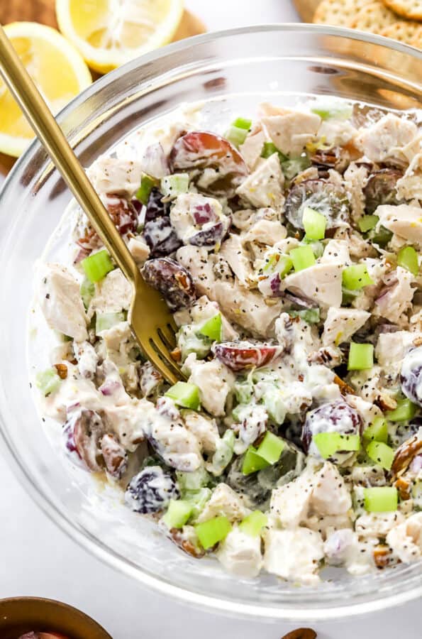 Chopped chicken with celery and grapes mixed in a creamy dressing in a bowl with a fork in the bowl with sliced lemon and crackers behind it.