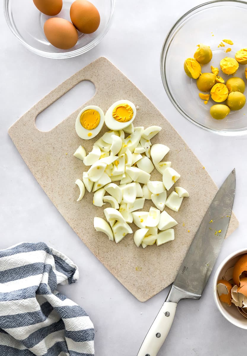 Cutting board wi chopped boiled eggs on it with more eggs in a bowl and egg yolks behind it with a knife on the board.