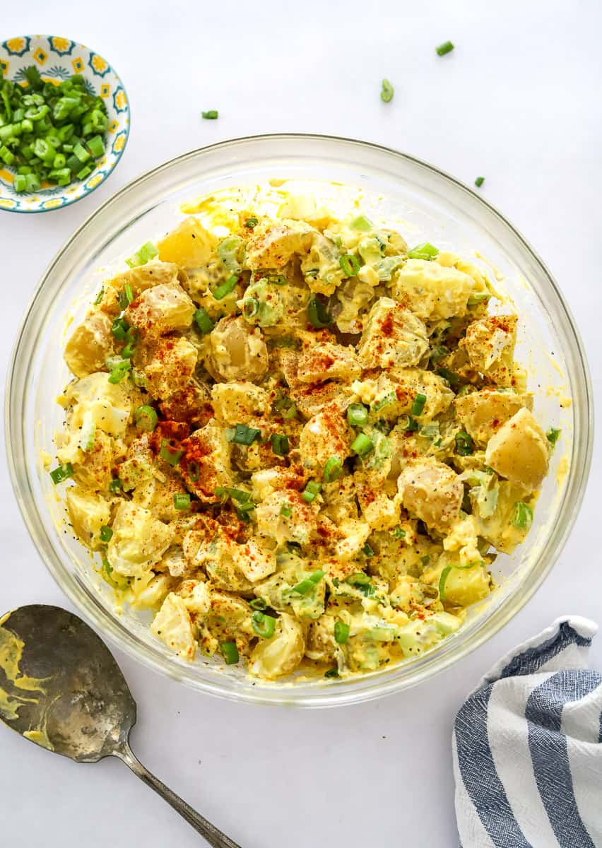 Assembled deviled egg potato salad in a mixing bowl with a silver serving spoon in front of it and chopped green onion in a bowl behind it.