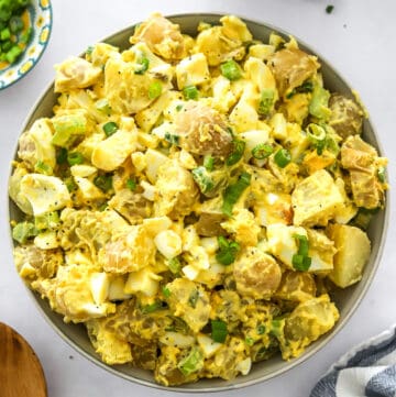 Bowl filled with potato salad with eggs and scallions in it with more scallions in a bowl behind it and a wood serving spoon and towel in front of it.