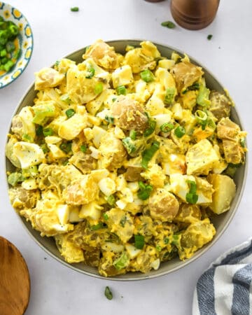 Bowl filled with potato salad with eggs and scallions in it with more scallions in a bowl behind it and a wood serving spoon and towel in front of it.