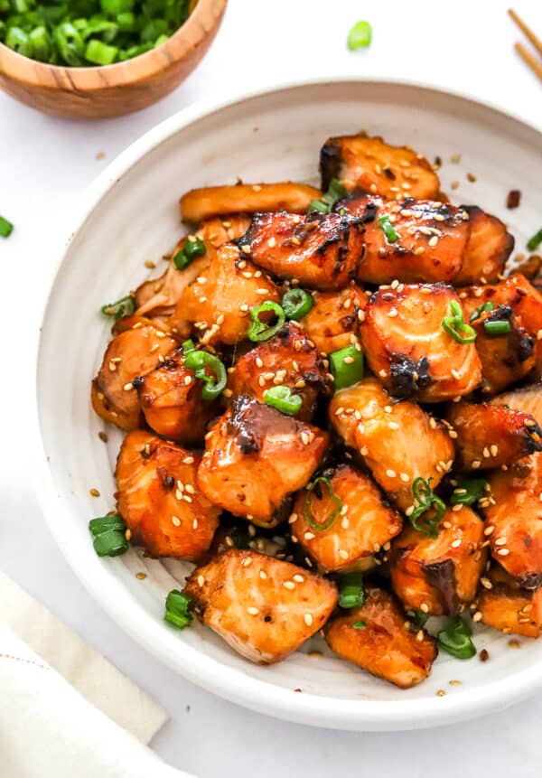 Cooked, salmon bites covered in teriyaki sauce on a plate with chopped scallions and sesame seeds on them.
