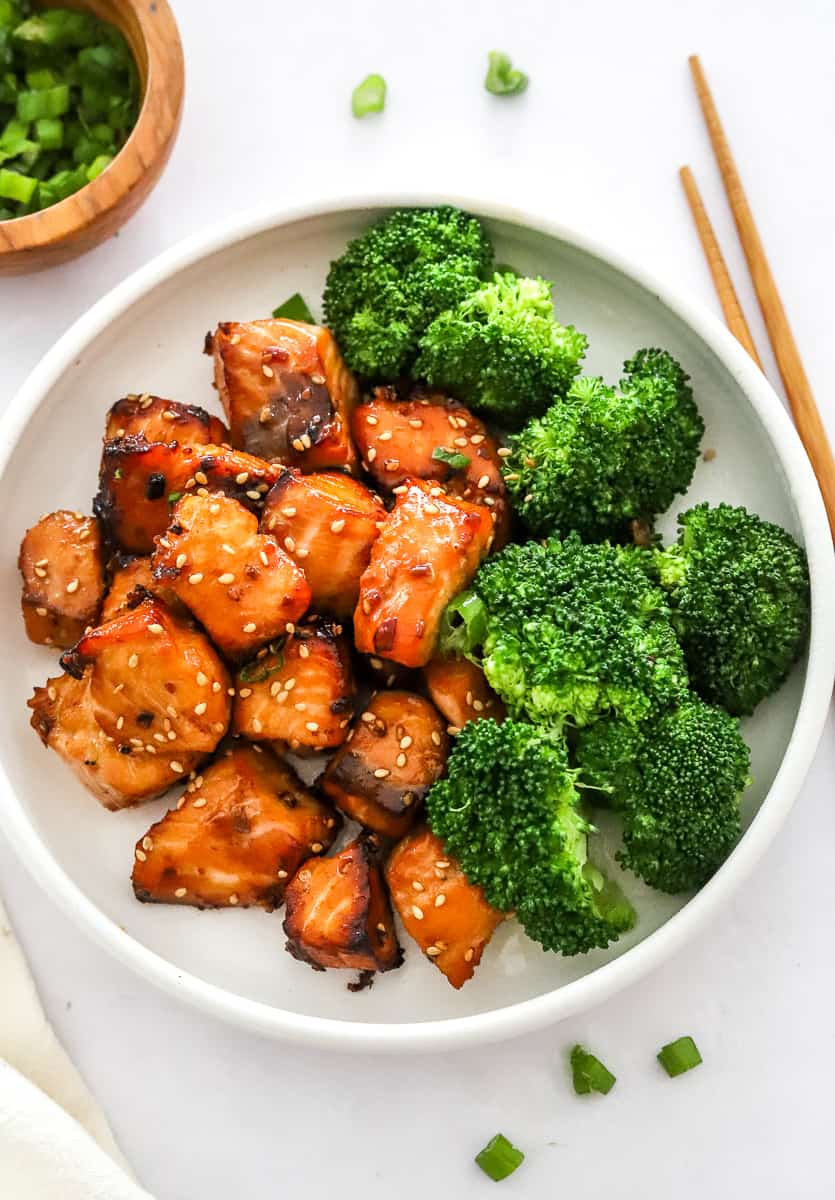 Teriyaki Air Fryer Salmon Bites cooked in a bowl with steamed broccoli with chopsticks and a bowl of green onion next to the bowl. 