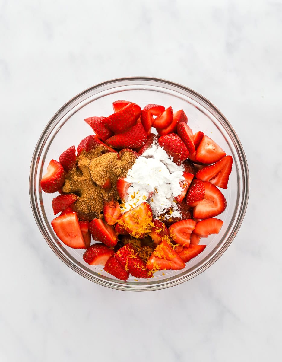 Sliced fresh strawberries with coconut sugar, starch and orange zest on them in a glass mixing bowl.